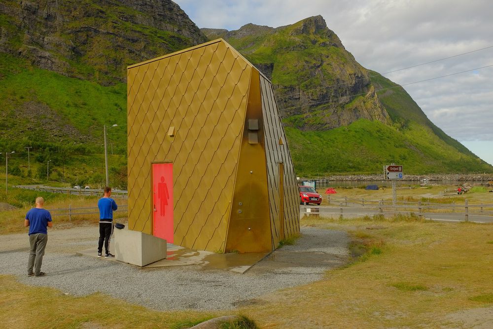 Two visitors standing next to the gold-clad toilet at Ersfjordstranda, mountains behind