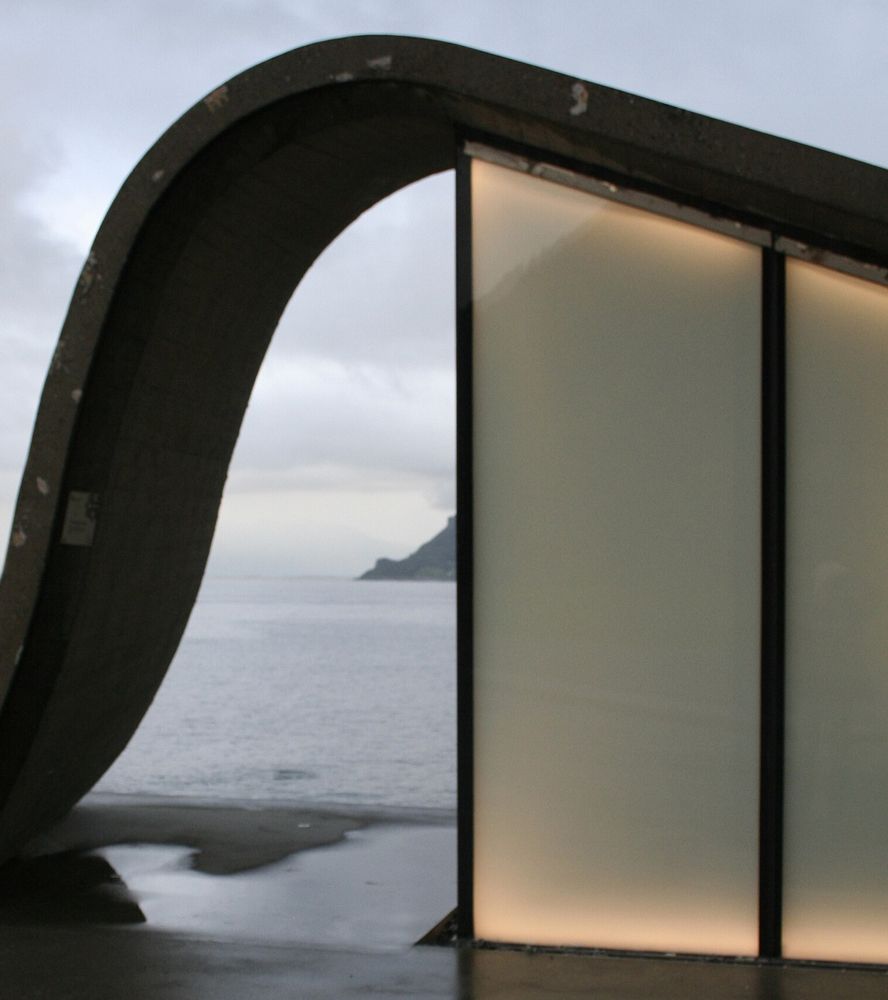 Ureddplassen rest stop: a curved concrete arch over a glowing glass toilet wall, with the sea behind